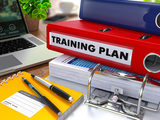 Red Ring Binder with Inscription Training Plan on Background of Working Table with Office Supplies, Laptop, Reports. Toned Illustration. Business Concept on Blurred Background. Mitch Gibson - Business Coach 53 Cook Parade 
